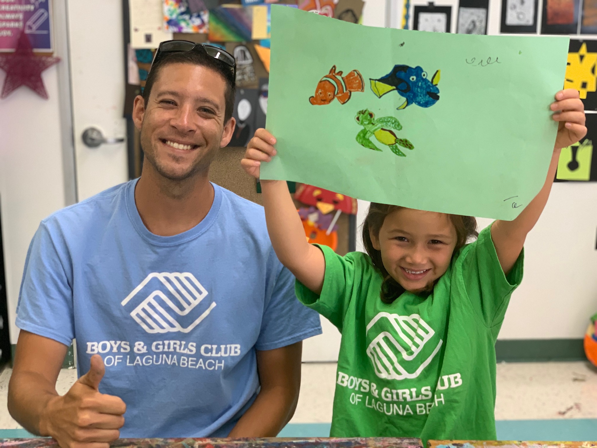thumbs up from male program volunteer with young girl showing off her artwork